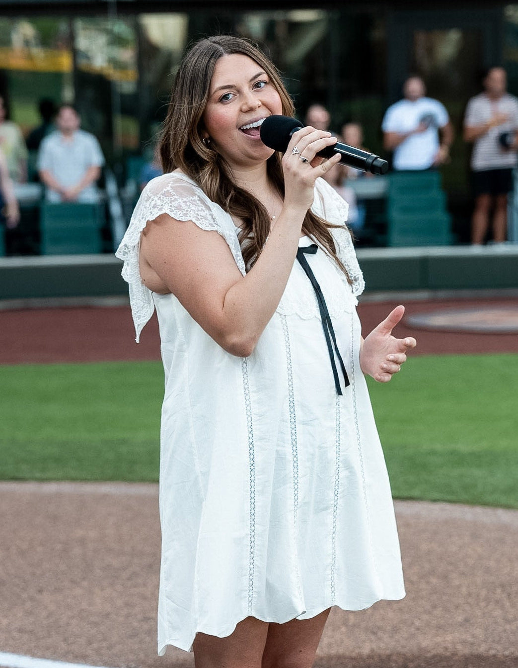 A woman stands on a baseball field holding a microphone, wearing a white sleeveless dress with lace trim and a black tie detail. The background shows the baseball field, stands, and spectators.